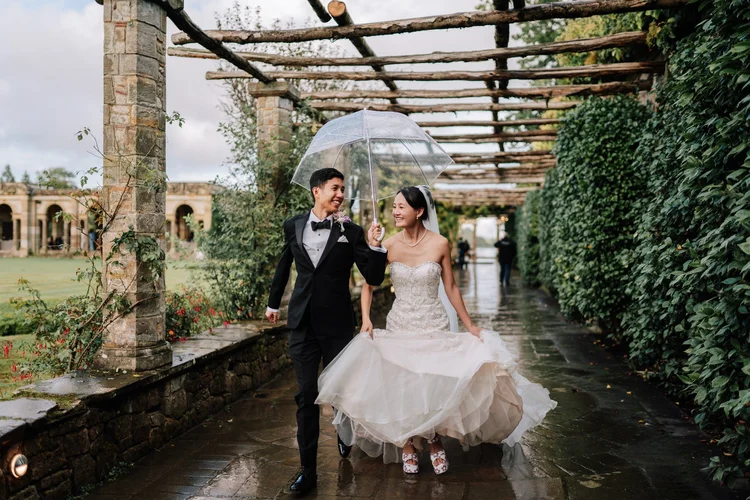 Bride and groom walking together under an umbrella through a stone pergola at their wedding venue